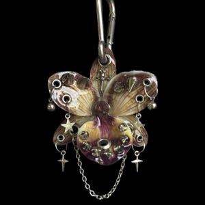May include: A decorative charm featuring a glossy, purple and cream-colored orchid flower adorned with silver-toned metal studs, small star-shaped charms, a tiny sword emblem, and a delicate silver chain, all attached to a metal carabiner clip.