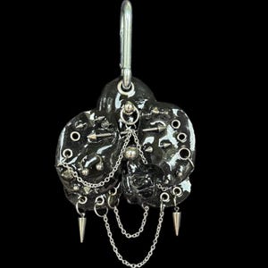 Puede incluir: Un dije negro brillante de múltiples lóbulos con herrajes de metal plateado, que incluyen pequeñas púas, anillos circulares y cadenas delicadas. La pieza está suspendida de un mosquetón plateado, creando un accesorio de aspecto industrial.