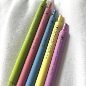 May include: An assortment of colorful pens in shades of green, pink, blue, yellow, and purple. Each pen has a different inspirational quote printed on it in white. The pens are arranged diagonally against a white background.