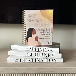 May include: A stack of books and a spiral-bound journal. The journal cover features a woman's profile and the text "SHE SPEAKS SCRIPTURE". Below, three white books read "HAPPINESS IS A JOURNEY" and "NOT THE DESTINATION".