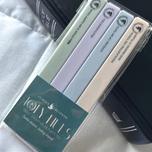 May include: A package of pastel-colored Bible study highlighters. The highlighters are in shades of green, purple, blue, and cream. Each highlighter has a different inspirational phrase printed on it. The package is labeled "Holy Hues" and "Clarke's Creations."