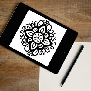 Könnte beinhalten: Ein schwarzes Tablet zeigt ein schwarz-weißes Blumenmuster. Ein schwarzer Bleistift und ein leeres weißes Papier liegen auf einer Holzoberfläche. Das Blumenmuster ist symmetrisch mit einer zentralen Blume und umgebenden Blättern und Wirbeln.