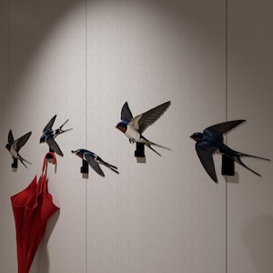 Op de afbeelding: Een set decoratieve wandhaken in de vorm van zwaluwen. De vogels zijn voornamelijk blauw, wit en zwart, met rode accenten. Een rode paraplu hangt aan een van de haken. De haken zijn op een witte muur gemonteerd.