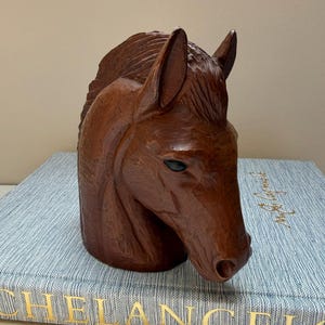 May include: A hand-carved brown wooden horse head sculpture featuring detailed mane textures, pointed ears, and dark eyes. The sculpture is positioned on top of two stacked books, one with the gold text MICHELANGELO and the other with IDAHO.