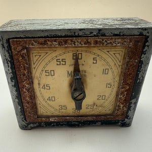May include: Vintage metal timer with a square, aged appearance. The face features a circular dial with numbers and a central hand. The casing is dark gray with rust-colored accents around the dial. The timer reads "Minutes".