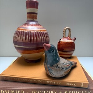 May include: A still life featuring a ceramic bird figurine with a red beak and a decorative vase with a striped neck. The bird sits atop a stack of books, including one titled "The Gardens of Winterthur."