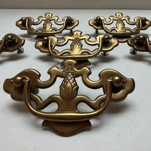 May include: A set of five brass drawer pulls featuring a decorative pineapple motif at the center of each backplate. Each pull includes a curved, swinging handle attached to a detailed metal plate with a warm, aged metallic finish and patina.