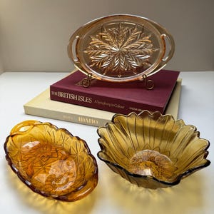 May include: An assortment of amber-colored glass serving dishes. The collection includes an oval tray with a floral design, a sunflower-shaped bowl, and a smaller, leaf-shaped dish. The pieces are arranged on a white surface.