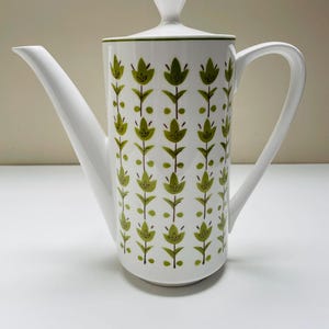 May include: White ceramic coffee pot with a long spout and handle. The pot is decorated with a repeating pattern of green stylized floral designs. The lid has a small knob and a green trim. The pot is on a white surface.