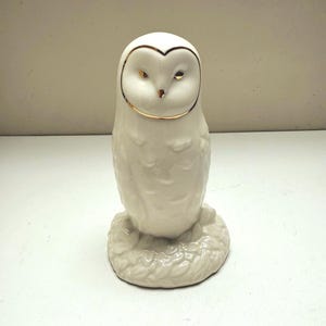 May include: A white ceramic owl figurine with gold accents around the eyes and head. The owl is perched on a textured base. The figurine has a smooth, glossy finish and is a decorative item.