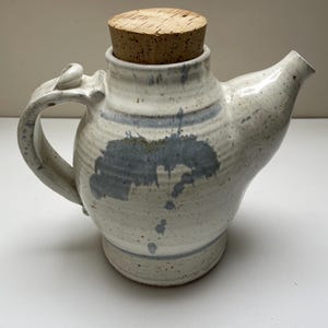 May include: Handmade ceramic teapot with a cork stopper. The teapot is off-white with blue-grey splatters and a matching handle. The spout is on the right side. The cork stopper is light brown. A unique piece for serving tea.