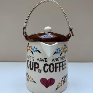 May include: A cream-colored ceramic coffee pot with a brown lid and a white wrapped handle. The body features hand-painted floral accents in blue, yellow, and green, a red heart, and the black text "LET'S HAVE ANOTHER CUP of COFFEE".