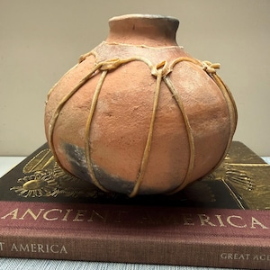 May include: A terracotta-colored, round vase with a narrow neck and a textured surface. The vase is adorned with a light-colored, woven design. It rests on a stack of books with the title "ANCIENT AMERICA".