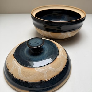 May include: Ceramic bowl and lid with a dark blue glaze and tan base. The lid features a small, round handle and a decorative white pattern. The bowl's interior is dark blue, and the rim is tan.
