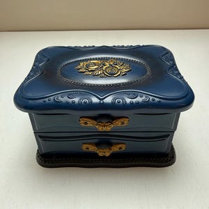 May include: A blue jewelry box with two drawers and a decorative top. The top features a gold-colored floral design. The box has gold-colored drawer pulls and a dark base. The jewelry box is a decorative storage item.