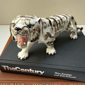 May include: A ceramic tiger figurine featuring a white base with brown stripes and a glossy finish. The tiger is depicted in a prowling stance with its mouth open, revealing a pink interior. The figurine is positioned on top of a black book.