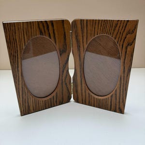 May include: A hinged, wooden double photo frame with an oak grain finish. Each frame has an oval opening for a photograph. The two frames are connected by a brass hinge, allowing them to stand open or fold closed.