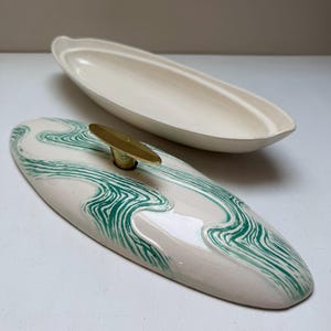 May include: An oval-shaped ceramic butter dish with a cream-colored base and a matching lid. The lid features a green marbled pattern and a gold-toned metal handle centered on top. The dish is separated into two pieces, showing the lid and base.