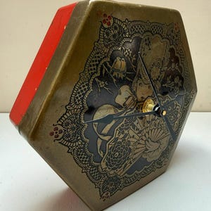 May include: A hexagonal wall clock with a metallic gold face featuring a black lace-like pattern, a central illustration of a person with a fan, and two small birds. The clock has black hands and a bright red side panel, creating a distinct contrast.