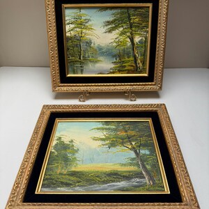 May include: Two framed landscape paintings. Each features a forest scene with a river or lake, painted in shades of green, blue, and yellow. The frames are gold with a black velvet matting. One painting is on a decorative stand.