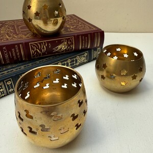 May include: Three spherical brass candle holders with cut-out designs. Two holders feature star-shaped cut-outs, while the front holder displays a pattern of small duck-shaped cut-outs. The items are arranged near two stacked hardcover books.