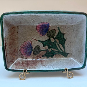 May include: Rectangular ceramic dish with a green rim and a thistle design. The dish has a speckled, off-white background with a brown and red mottled pattern. The thistle flowers are purple, and the leaves are green.