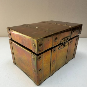 May include: A rectangular brass storage chest with a hinged lid and a metallic finish. The box features reinforced corners, vertical and horizontal metal bands, and a central latch mechanism on the front. The surface displays a warm, iridescent patina.