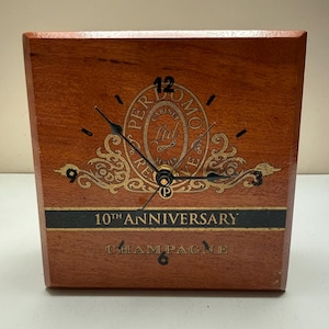 May include: A wooden clock with a rectangular shape. The clock face features a gold-colored decorative design with the text "PERLOMO CABINET REIMS Ltd." and "10TH ANNIVERSARY CHAMPAGNE". The clock hands are black.