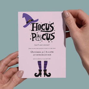 Könnte beinhalten: Eine rosa Einladungskarte mit den Worten "Hocus Pocus" in Schwarz, einem Hexenhut, einem Kürbis und Hexenbeinen. Der Text "Let's get spooky!" und Veranstaltungsdetails sind ebenfalls enthalten. Die Karte wird von zwei Händen gehalten.