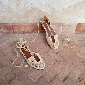 Könnte beinhalten: Ein Paar beige Häkelschuhe mit braunen Innensohlen und langen, dünnen Schnürsenkeln. Die Schuhe stehen auf einem roten Ziegelboden, mit einer strukturierten, cremefarbenen Wand im Hintergrund. Die Schuhe haben ein gewebtes Design.