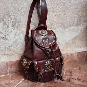 Può includere: Uno zaino in pelle marrone con borchie e abbellimenti metallici decorativi. Lo zaino presenta una chiusura con patta, tasche laterali e spallacci regolabili. La borsa ha una chiusura con coulisse e dettagli con fibbia.