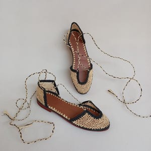 Könnte beinhalten: Ein Paar geflochtener Raffia-Ballerinas mit schwarzem Besatz und braunen Lederinnensohlen. Die Schuhe haben ein Schnürdesign mit schwarz-weiß gestreiften Bändern. Die Sohlen sind rot.