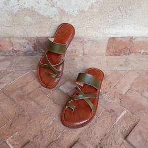 Könnte beinhalten: Ein Paar braune Ledersandalen mit olivgrünen Riemen. Die Sandalen haben eine Zehenschlaufe und gekreuzte Riemen über dem Fuß. Die Sandalen werden auf einer Ziegeloberfläche präsentiert.