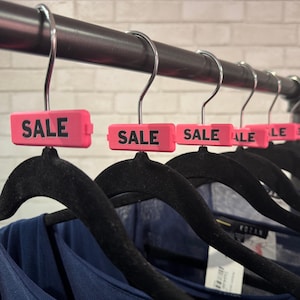 May include: A row of black velvet-textured clothes hangers suspended from a metal rod. Each hanger features a bright pink rectangular plastic tag attached to the hook, with the word SALE printed in bold black capital letters on the front.