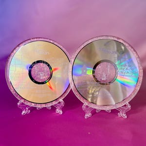 Könnte beinhalten: Zwei dekorative CDs mit rosafarbenen Glitzerharz-Rändern. Die CDs werden auf transparenten Kunststoffständern vor einem violett-rosa Farbverlaufshintergrund präsentiert. Eine CD hat Text und die andere eine Regenbogenreflexion.