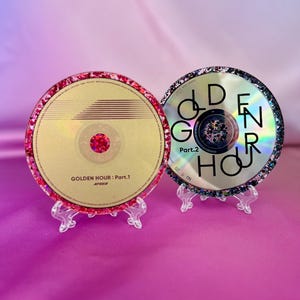 Könnte beinhalten: Zwei dekorative CDs, jeweils auf einem transparenten Ständer präsentiert. Eine CD hat eine goldene Oberfläche mit dem Text "GOLDEN HOUR: Part.1" und einen roten Glitzerrahmen. Die andere CD hat eine silberne Oberfläche mit dem Text "GOLDEN HOUR" und einen schwarzen Glitzerrahmen.