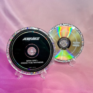 Könnte beinhalten: Zwei CDs mit glitzerbedeckten Rändern und transparenten Ständern. Eine CD ist schwarz mit weißem Text und den Worten "SPIN OFF: FROM THE WITNESS". Die andere CD ist goldfarben mit Regenbogen-Schimmer. Beide haben einen zentralen Schmuckstein.