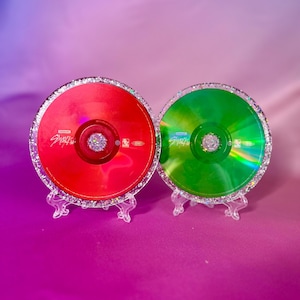 Puede incluir: Dos CD decorativos, uno rojo y otro verde, cada uno con un borde de purpurina plateada brillante y una joya central. Los CD se exhiben sobre soportes de acrílico transparente, sobre un fondo degradado púrpura y rosa.