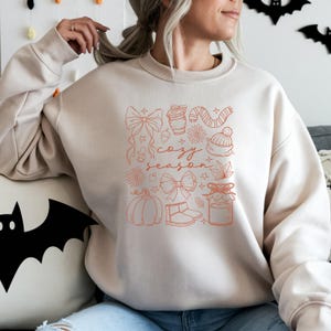 May include: A beige sweatshirt with a fall-themed design in orange. The design includes a pumpkin, bows, a coffee cup, a candy cane, a hat, and the words "cozy season."