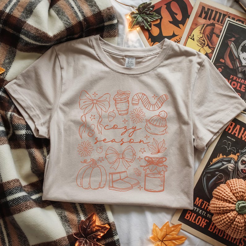 May include: A beige t-shirt with a red graphic design featuring the words "cozy season" surrounded by fall-themed illustrations, including a pumpkin, bow, and a cup of coffee. The shirt is displayed on a plaid blanket.