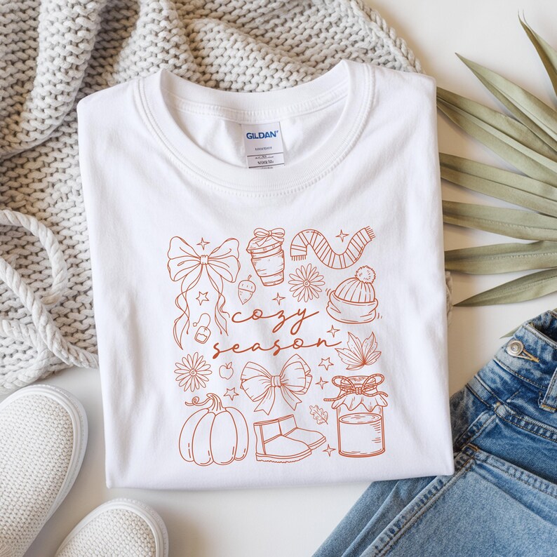 May include: White t-shirt with a "Cozy Season" design in brown. The design includes a pumpkin, bow, coffee cup, scarf, hat, and other fall-themed illustrations. The shirt is folded on a white surface with a neutral-colored blanket and jeans in the background.