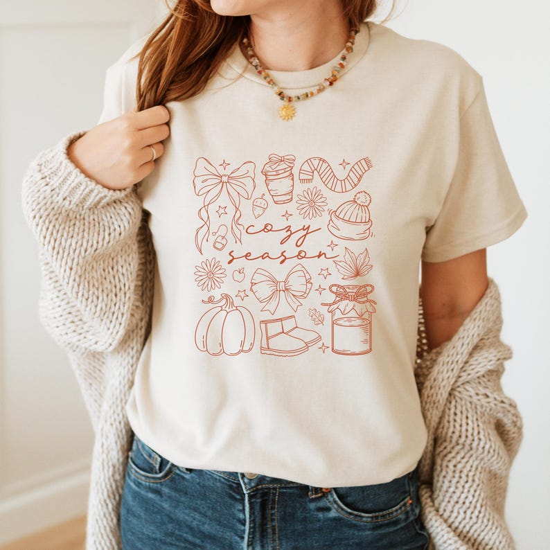 May include: Beige t-shirt with a red outline design featuring bows, a coffee cup, a scarf, a beanie, leaves, a pumpkin, boots, and a jar. The words "cozy season" are written in cursive script.