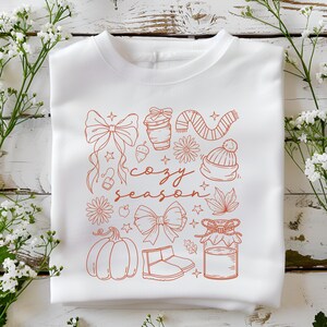May include: White sweatshirt with a rust-colored graphic featuring bows, a coffee cup, a scarf, a beanie, a pumpkin, boots, and the words "cozy season". The design includes stars, flowers, and leaves.