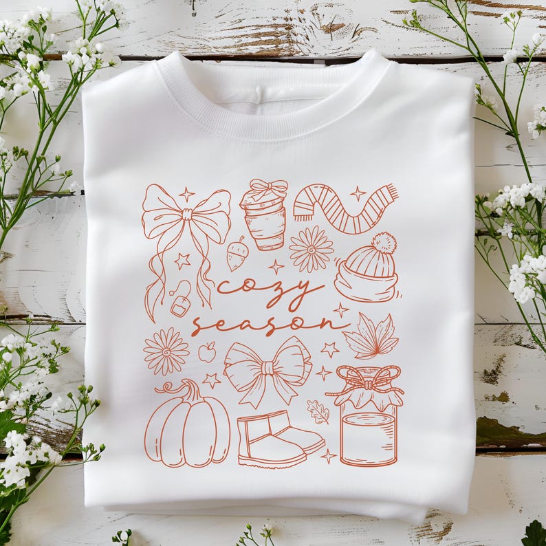 May include: White sweatshirt with a rust-colored graphic featuring bows, a coffee cup, a scarf, a beanie, a pumpkin, boots, and the words "cozy season". The design includes stars, flowers, and leaves.