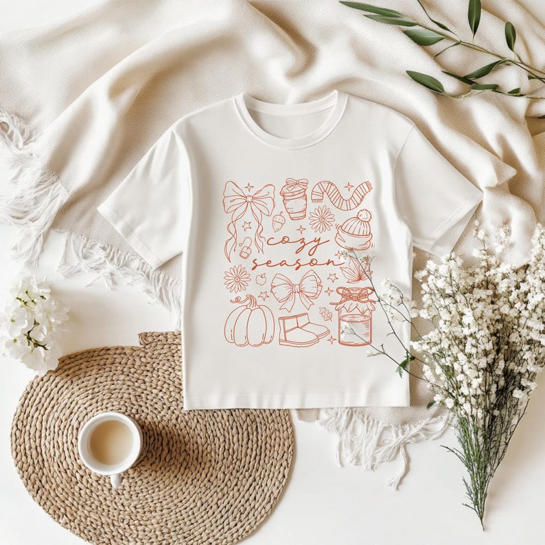 May include: White t-shirt with a crew neck featuring a red illustration of fall-themed elements, including bows, a coffee cup, candy canes, a hat, flowers, a pumpkin, and the words "cozy season". The shirt is displayed on a white surface.