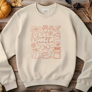 May include: Cream-colored sweatshirt with a fall-themed graphic in orange. The design includes a pumpkin, bows, a coffee cup, a scarf, a hat, and the words "cozy season."