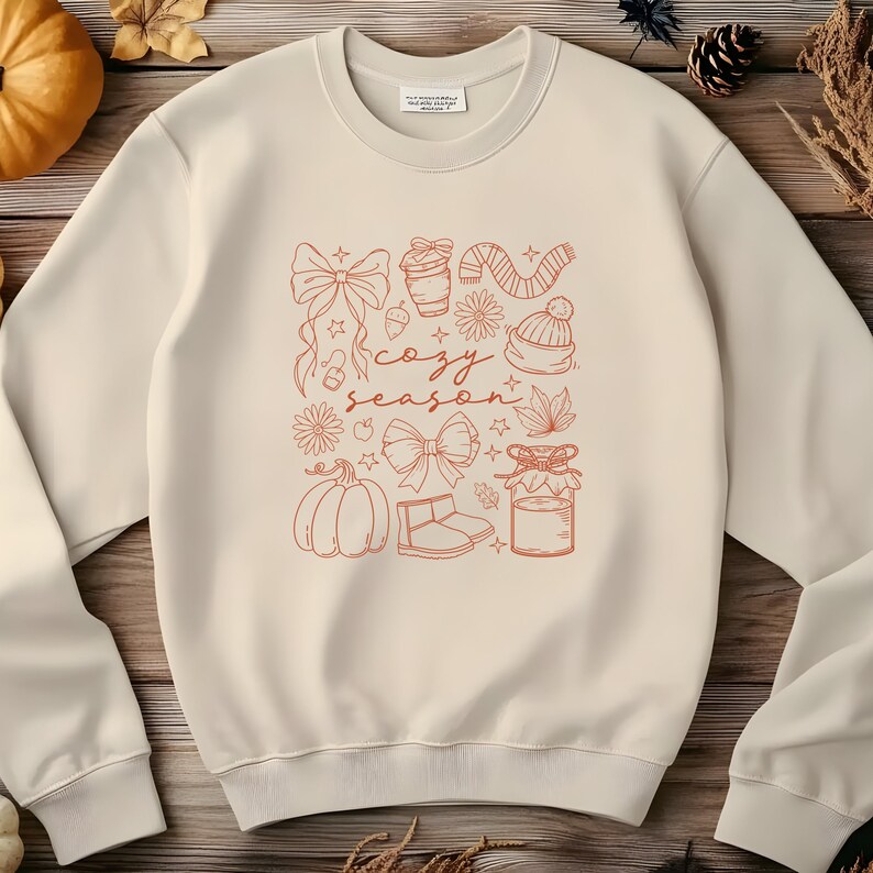 May include: Cream-colored sweatshirt with a fall-themed graphic in orange. The design includes a pumpkin, bows, a coffee cup, a scarf, a hat, and the words "cozy season."