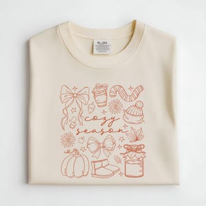 May include: Cream-colored sweatshirt with a rust-colored graphic. The graphic includes a bow, coffee cup, scarf, hat, flowers, pumpkin, boots, and the words "cozy season".