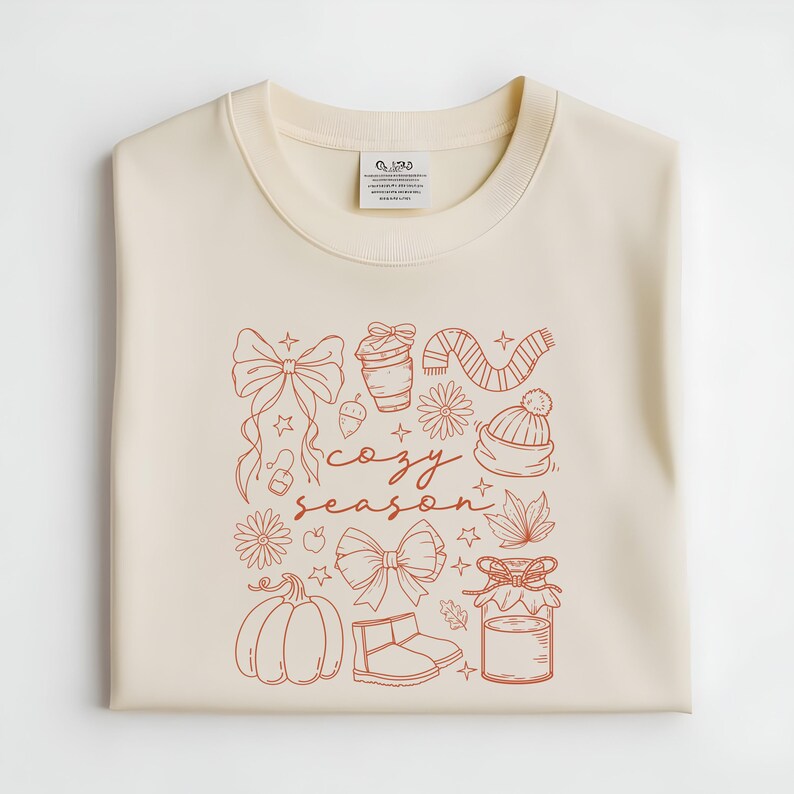 May include: Cream-colored sweatshirt with a rust-colored graphic. The graphic includes a bow, coffee cup, scarf, hat, flowers, pumpkin, boots, and the words "cozy season".