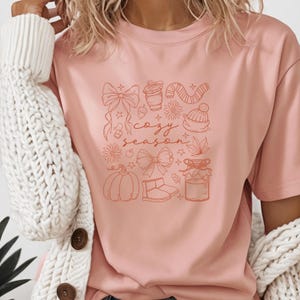 May include: A light pink t-shirt featuring a collection of fall-themed illustrations in a burnt orange color. The design includes bows, a coffee cup, a scarf, a beanie, a pumpkin, and the words "cozy season".
